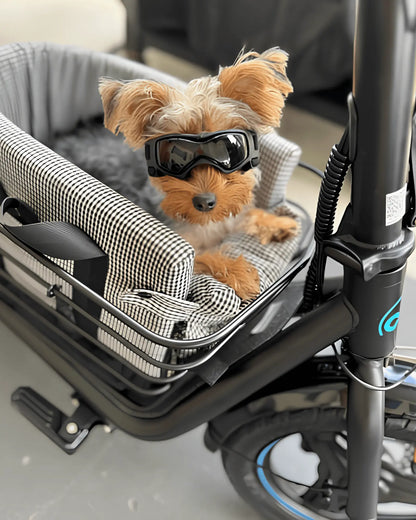 Small dog wearing goggles sitting in a pet carrier with a padded basket insert for pets with bow design, gray color, and checkered lining
